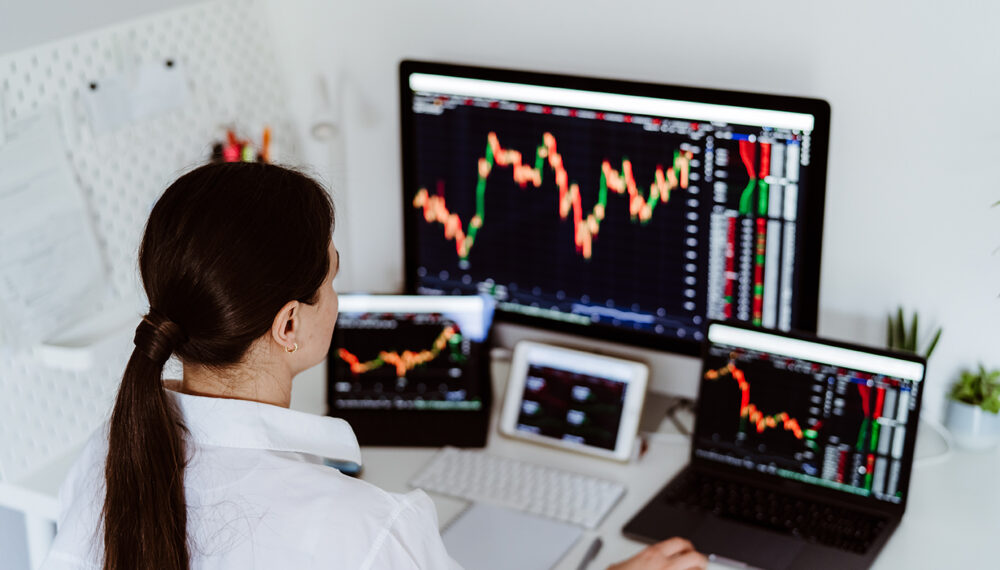 Back view of young female cryptocurrency trader sitting at desk looking at multiple screens monitoring stock financial market. Business woman investor analyst analyze currency growth graphs and charts.